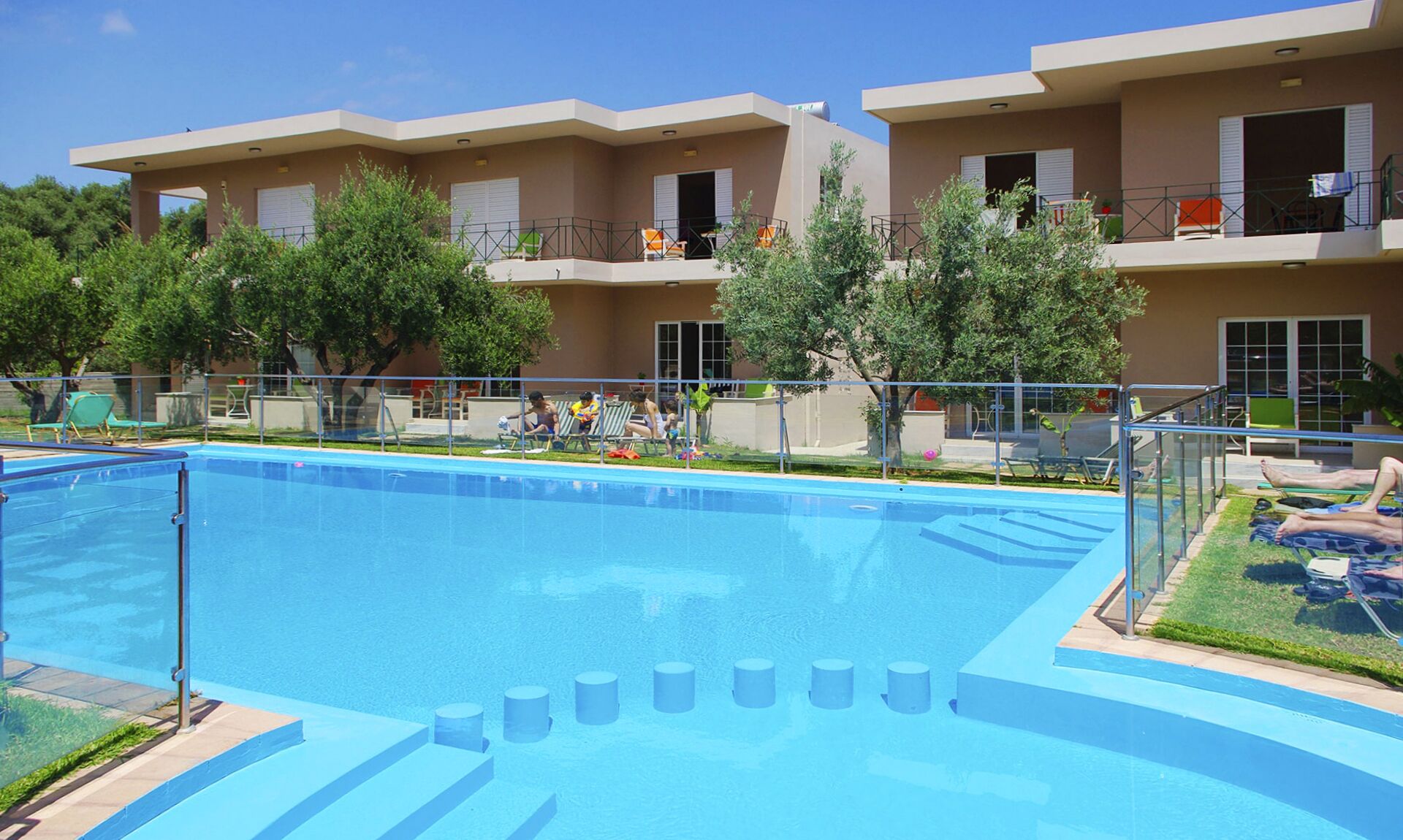 Una piscina con escalones de acceso, rodeada por una valla. Hay sillas de descanso y sombrillas junto a la piscina.
