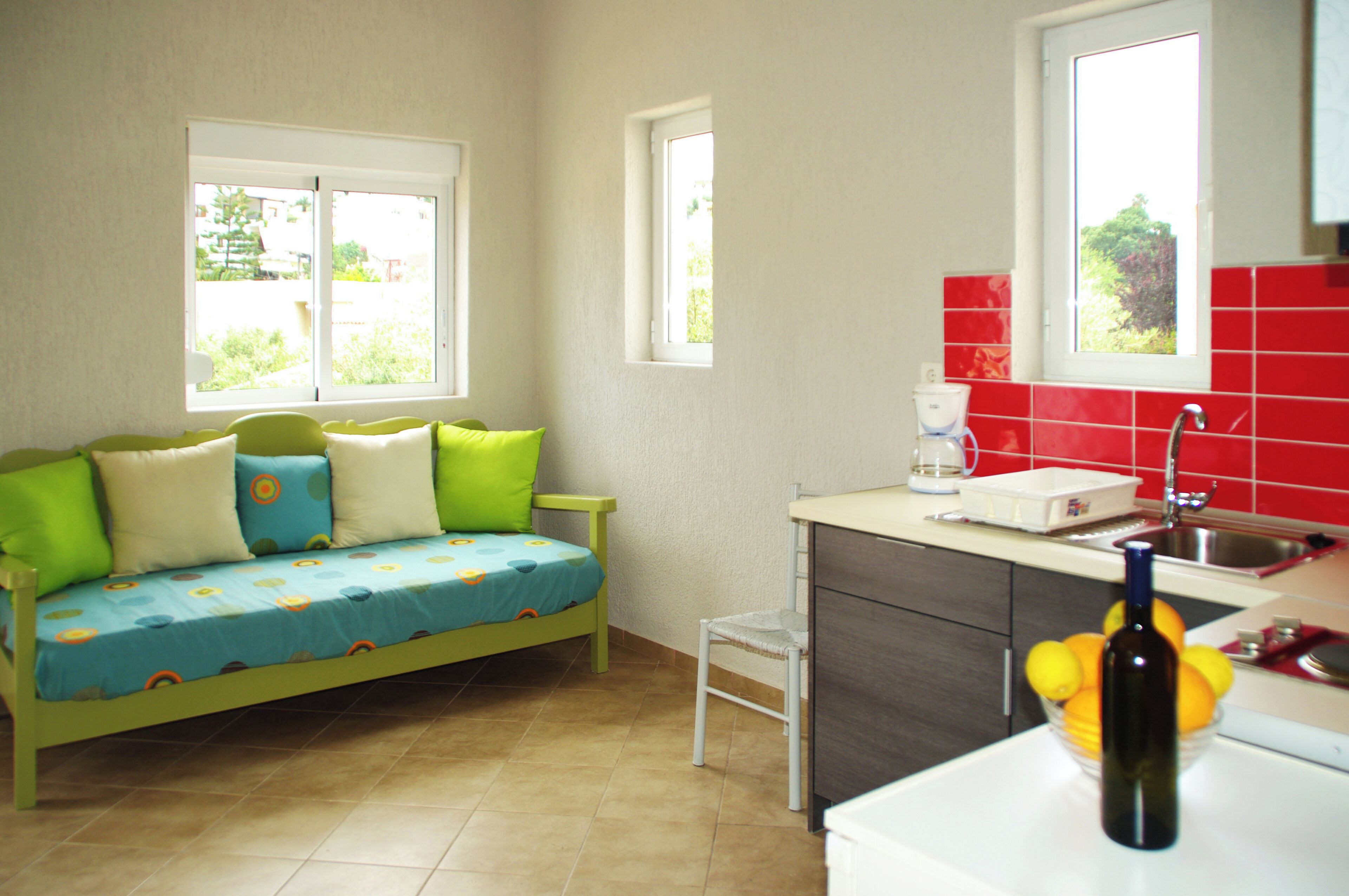 Una habitación pequeña y luminosa con un sofá verde, una zona de cocina con baldosas rojas y una ventana que proporciona luz natural.
