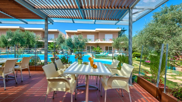 A covered outdoor seating area with a pool view, white chairs, and a table with a drink. - Elea apartments (Chania)