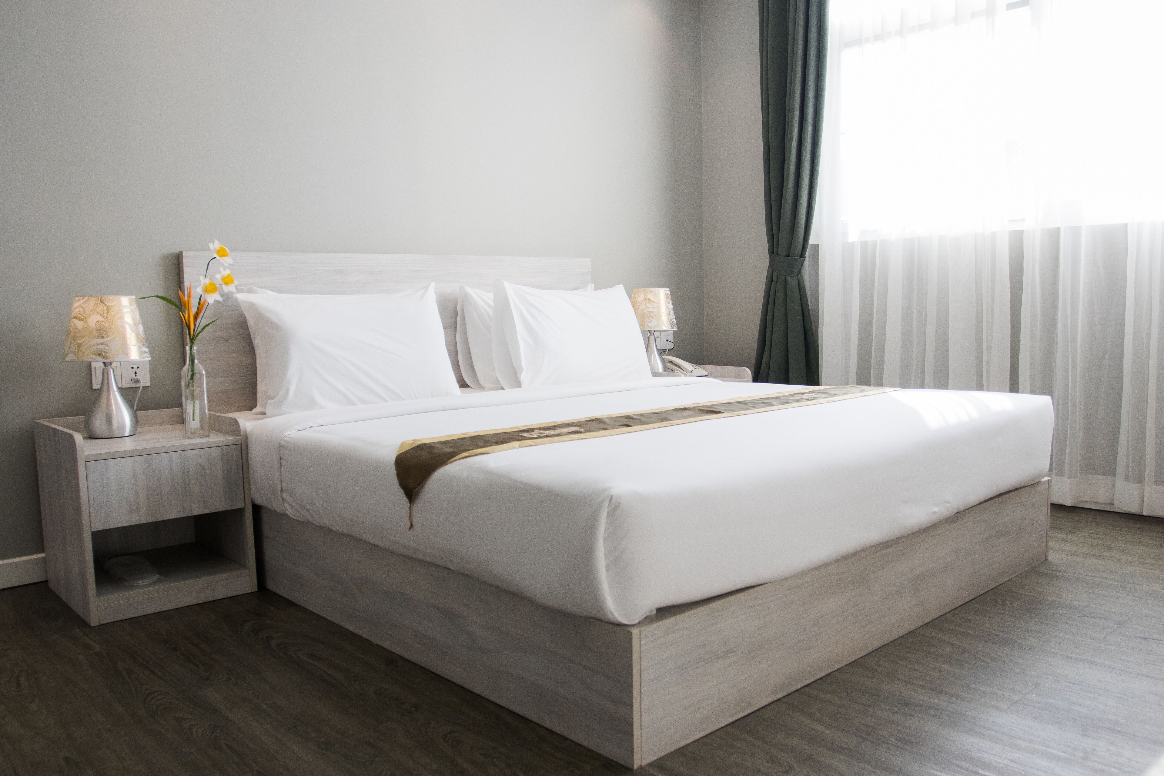 A modern hotel room with a large bed, two bedside tables, a vase with flowers, and a window with curtains.