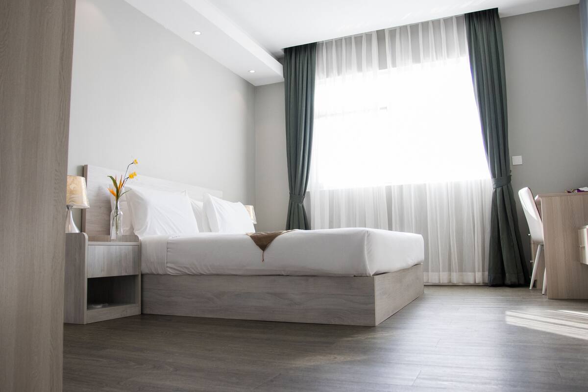 Ein modernes Schlafzimmer mit einem großen Bett, einem Nachttisch mit Lampe und Blick ins Freie durch transparente Vorhänge.