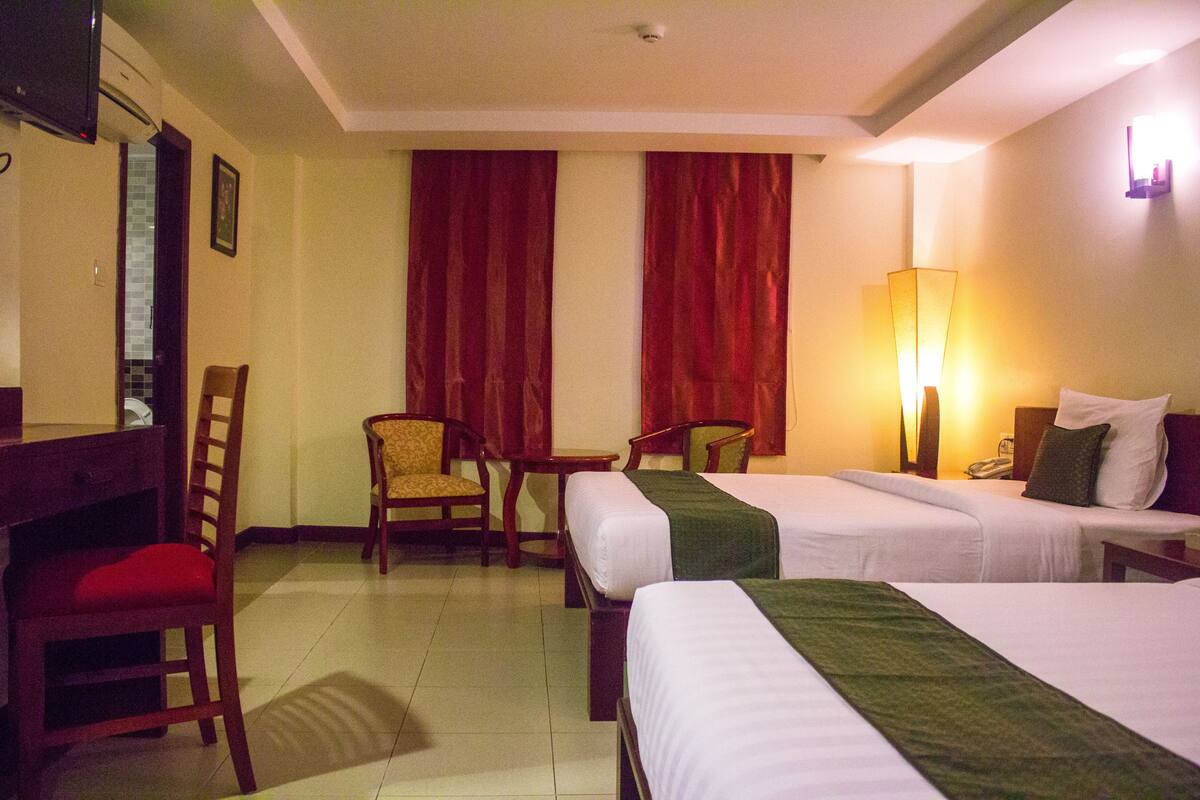 A hotel room with two beds, a desk, a chair, and a lamp.