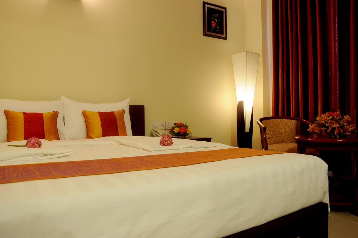A hotel room with a large bed, two pillows, a nightstand with a lamp, a chair, and a vase of flowers.