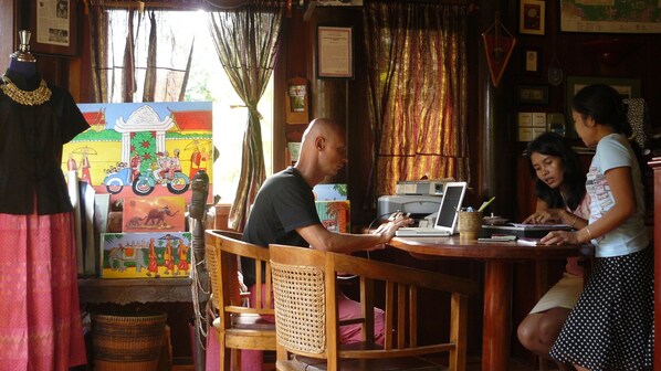 A room with a wooden table, a laptop, and a person working. There is a colorful mural on the wall and a mannequin with a necklace. - Terres Rouges Lodge (Ban Lung)