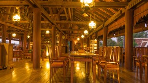 Un amplio comedor interior con mesas y sillas de madera, pilares grandes y lámparas colgantes.