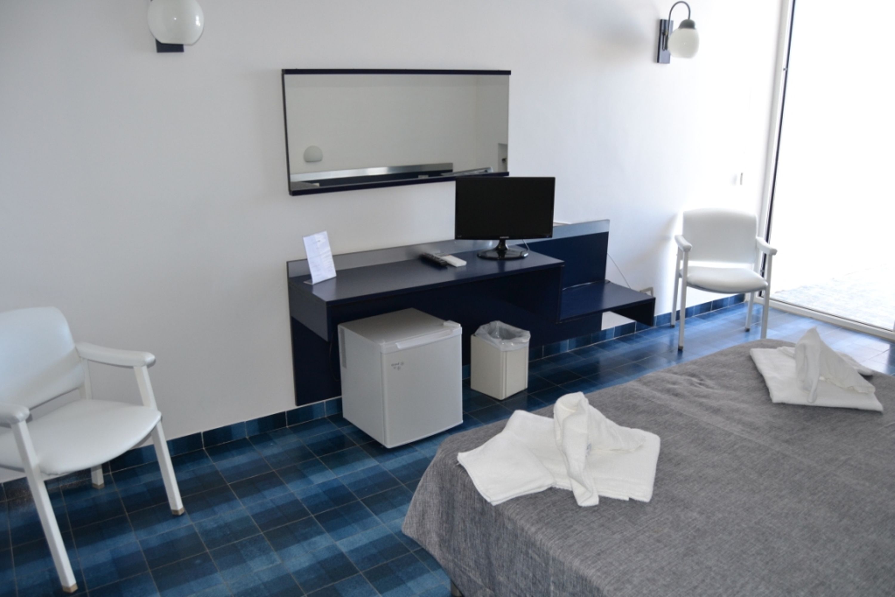 Ein Hotelzimmer mit einem Bett, einem Schreibtisch, einem Stuhl, einem Mülleimer und einem kleinen Kühlschrank.
