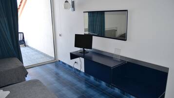 Una moderna camera d'hotel con una televisione a schermo piatto su una consolle blu scuro, uno specchio grande sopra e vista sulla zona esterna attraverso una porta in vetro.