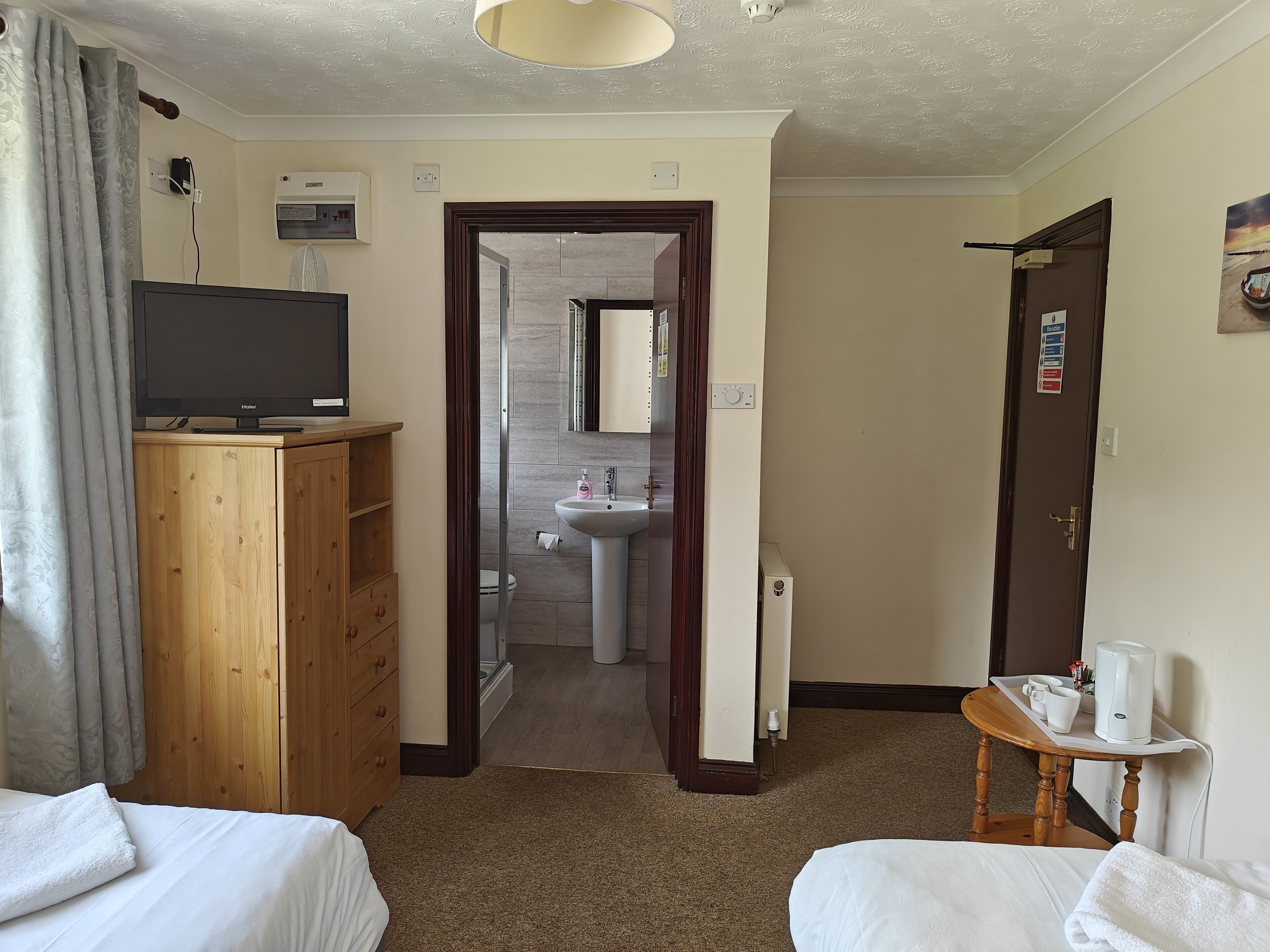 Habitación de hotel con armario de madera, televisor, cama con toalla blanca, mesita con cafetera y baño visible a través de una puerta abierta.