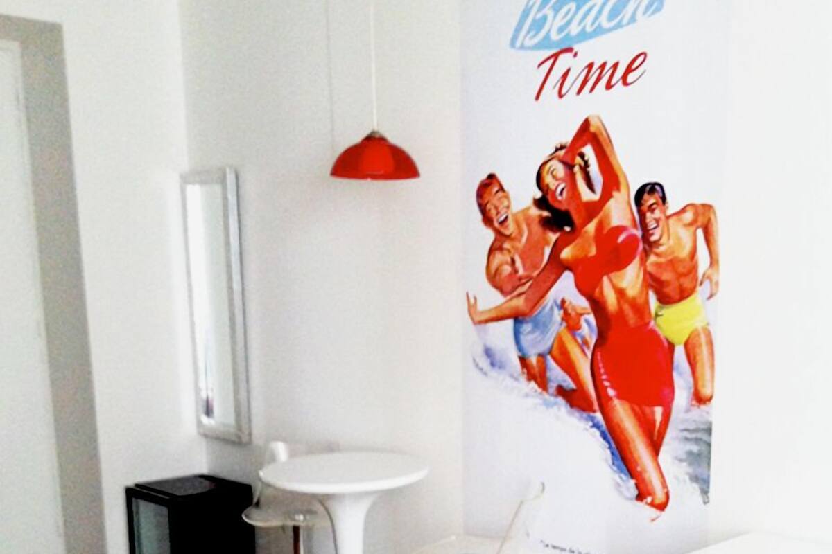 A modern hotel room with a wooden floor, a red carpet, a white sink, two bar stools, a red pendant light, and a poster of beach-themed artwork.