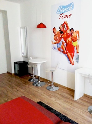 Una habitación de hotel moderna con piso de madera, alfombra roja, lavamanos blanco, dos taburetes altos, una lámpara colgante roja y un cartel con arte temático de playa.