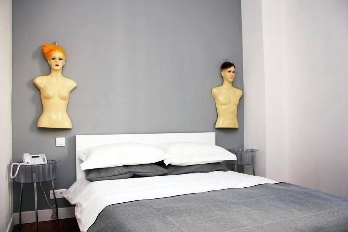 A bedroom with a bed, white pillows, a bedside table with a lamp, and two wall-mounted mannequin heads.