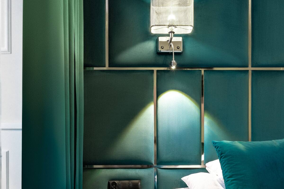 A modern bedroom with teal walls, a bed with white and teal bedding, a nightstand with a lamp, and a drawer unit.