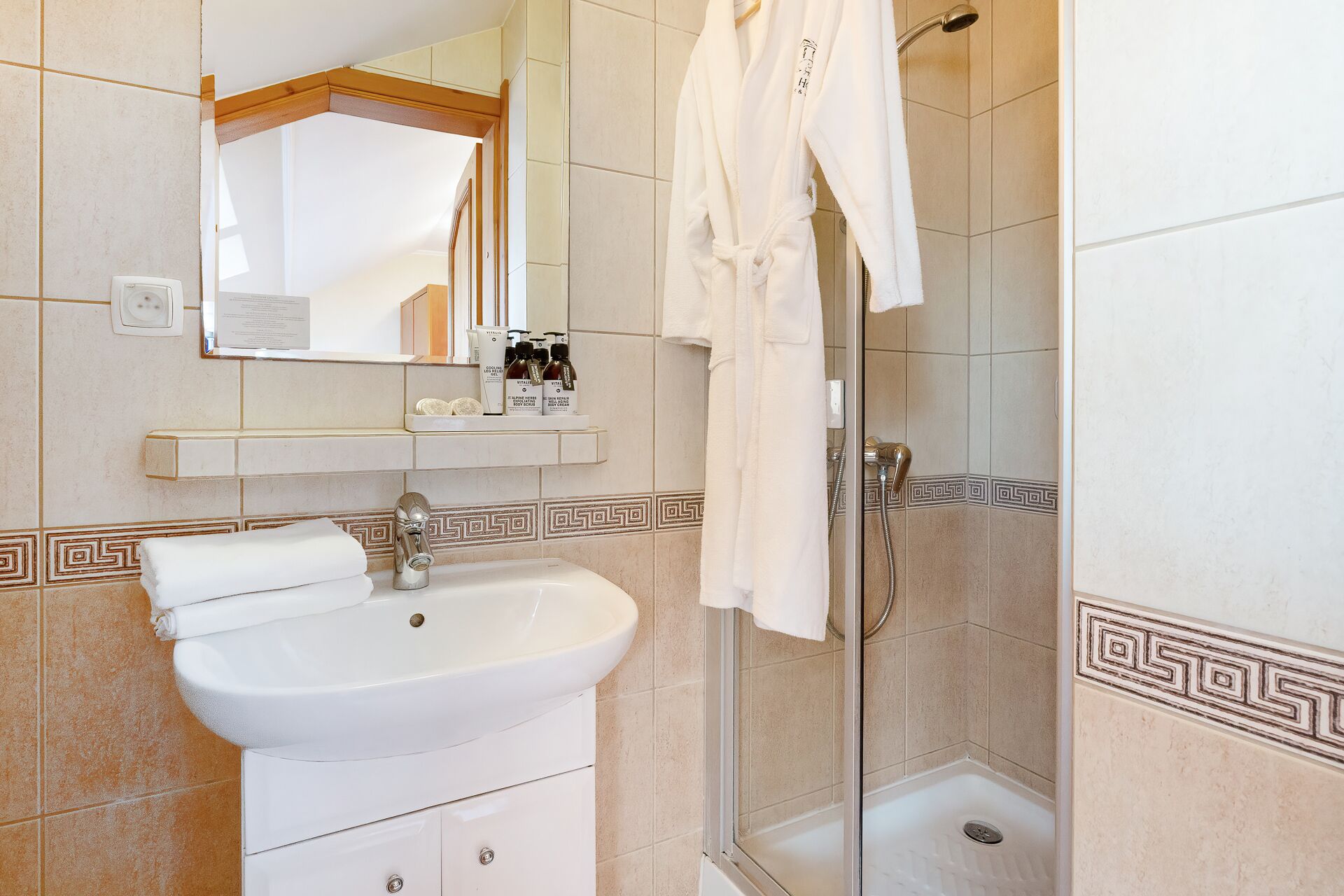 Ein Badezimmer mit einem weißen Waschbecken, einem Spiegel und einer Dusche. Auf dem Regal über dem Waschbecken liegen Toilettenartikel.