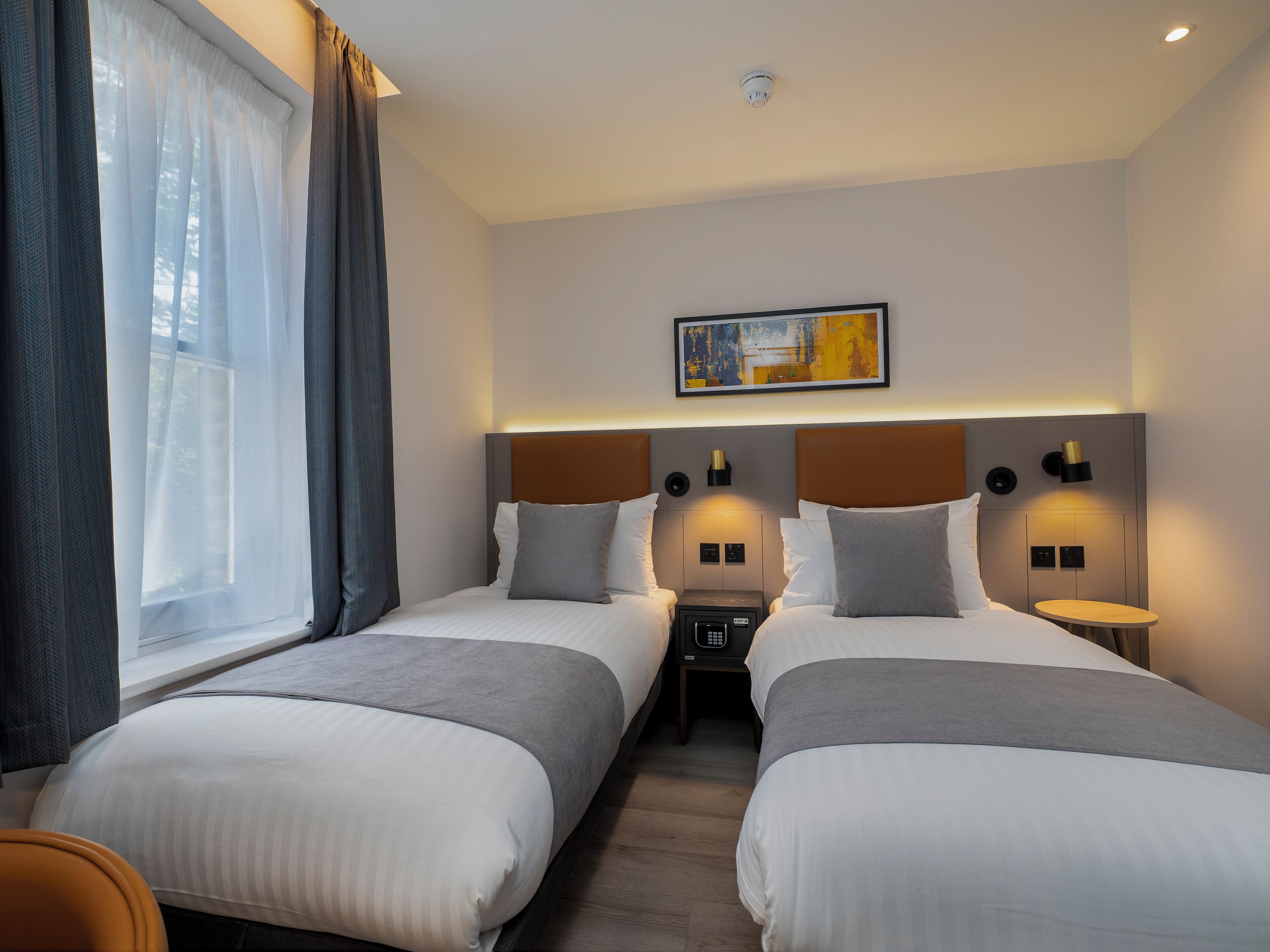 Habitación de hotel con dos camas, una mesita de noche con lámpara, un cuadro en la pared y una ventana con cortinas.