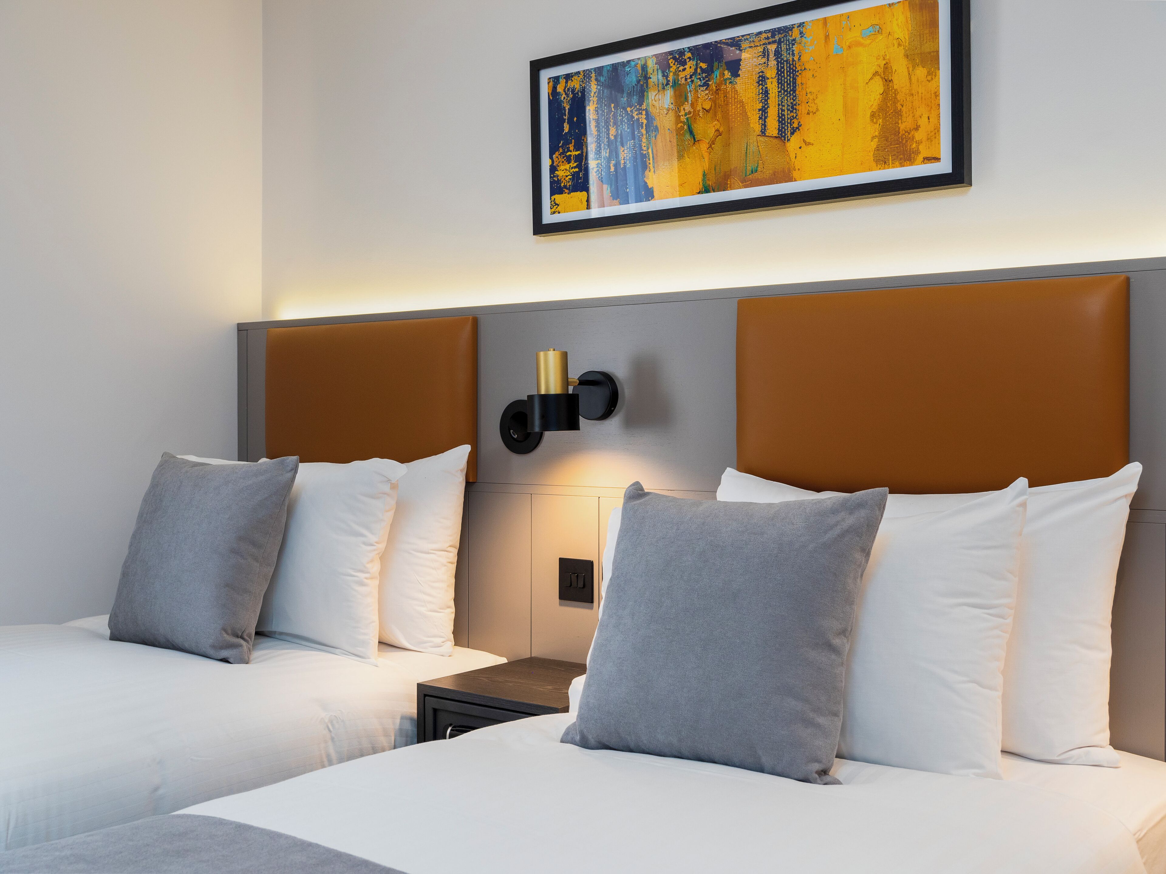 Une chambre d’hôtel avec deux lits, une tête de lit dotée d’un luminaire, un tableau abstrait encadré et une petite table de chevet.