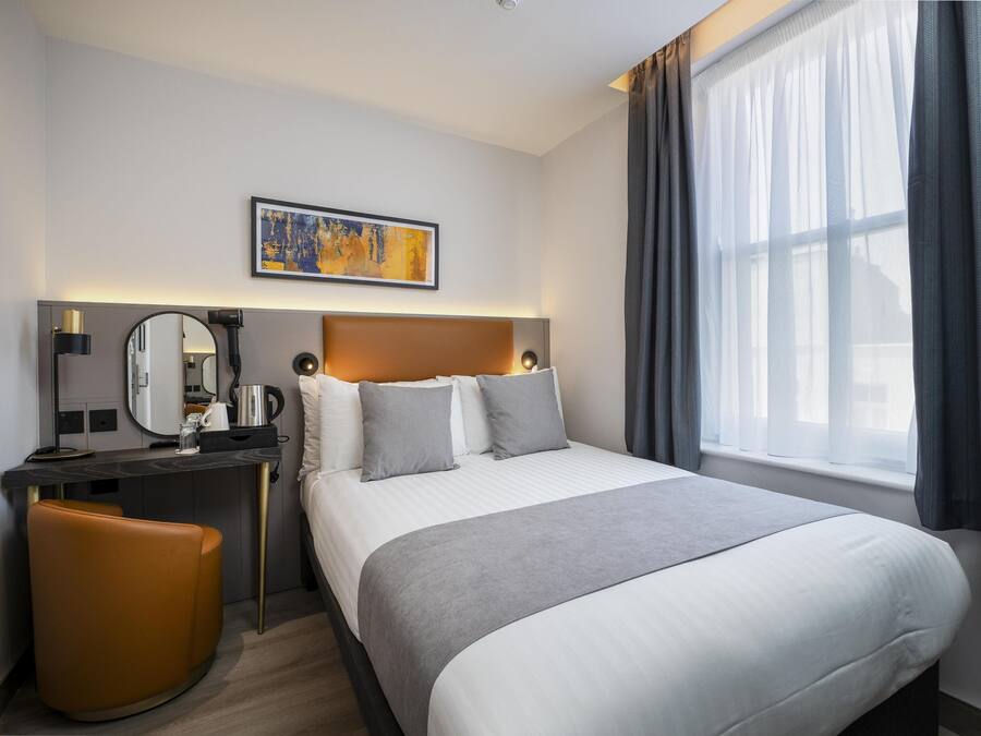 A hotel room with a bed, a desk, a chair, a mirror, and a painting on the wall.