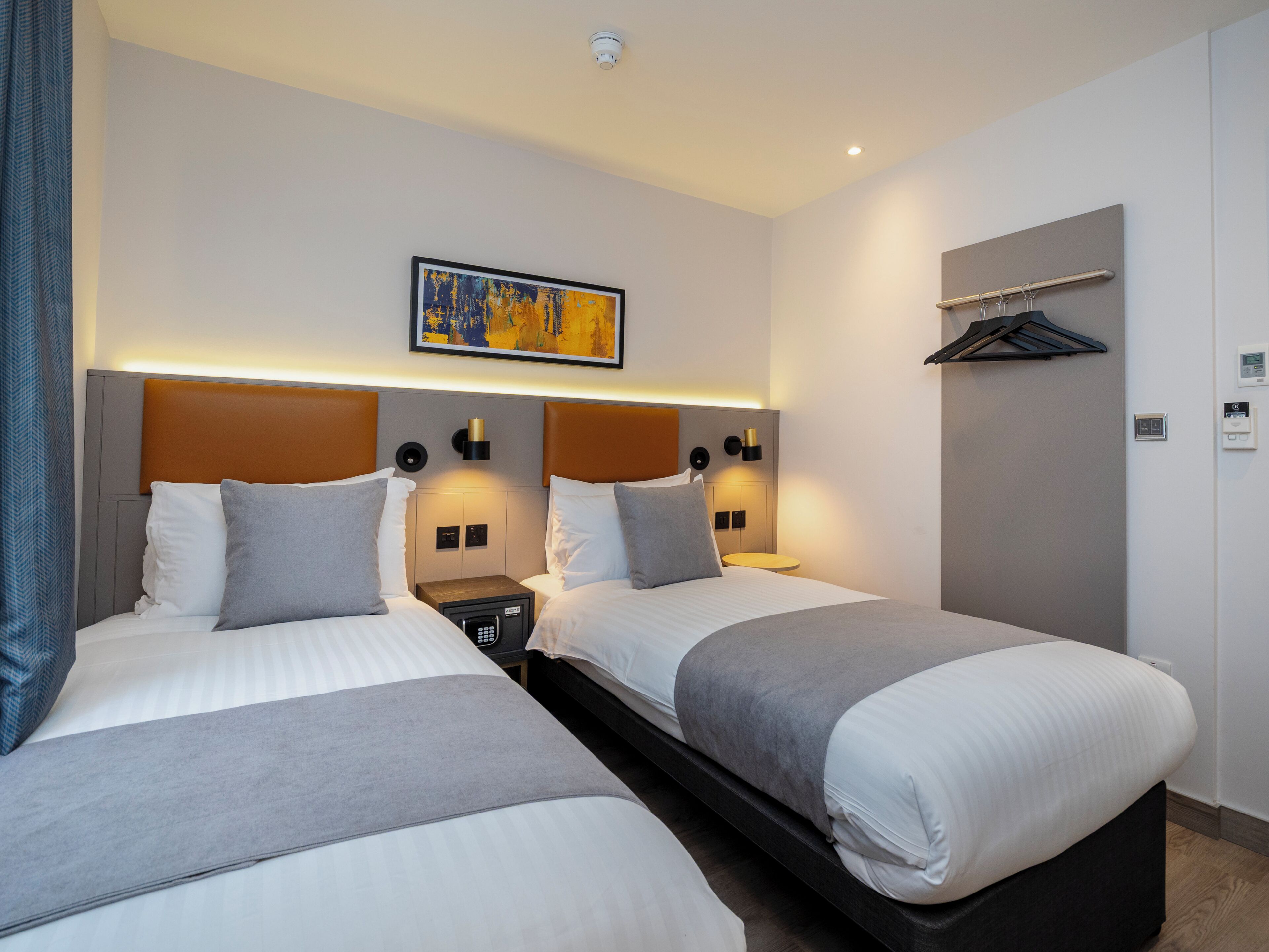 A hotel room with two beds, a headboard with integrated lighting, a bedside table with a lamp, a wall-mounted artwork, and a coat rack.