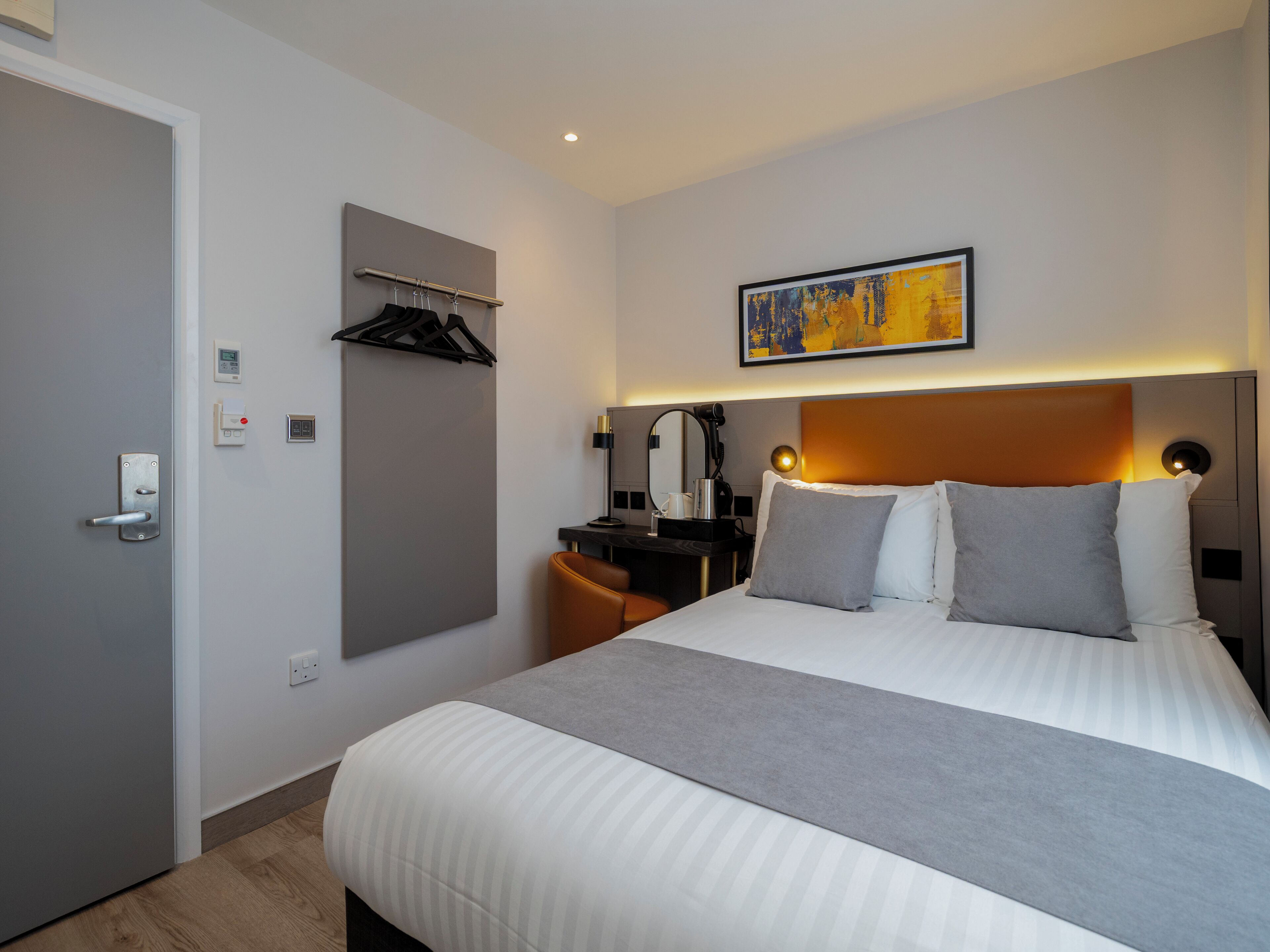 Ett hotellrum med en säng, ett skrivbord, en stol, en klädställning och en tavla på väggen.