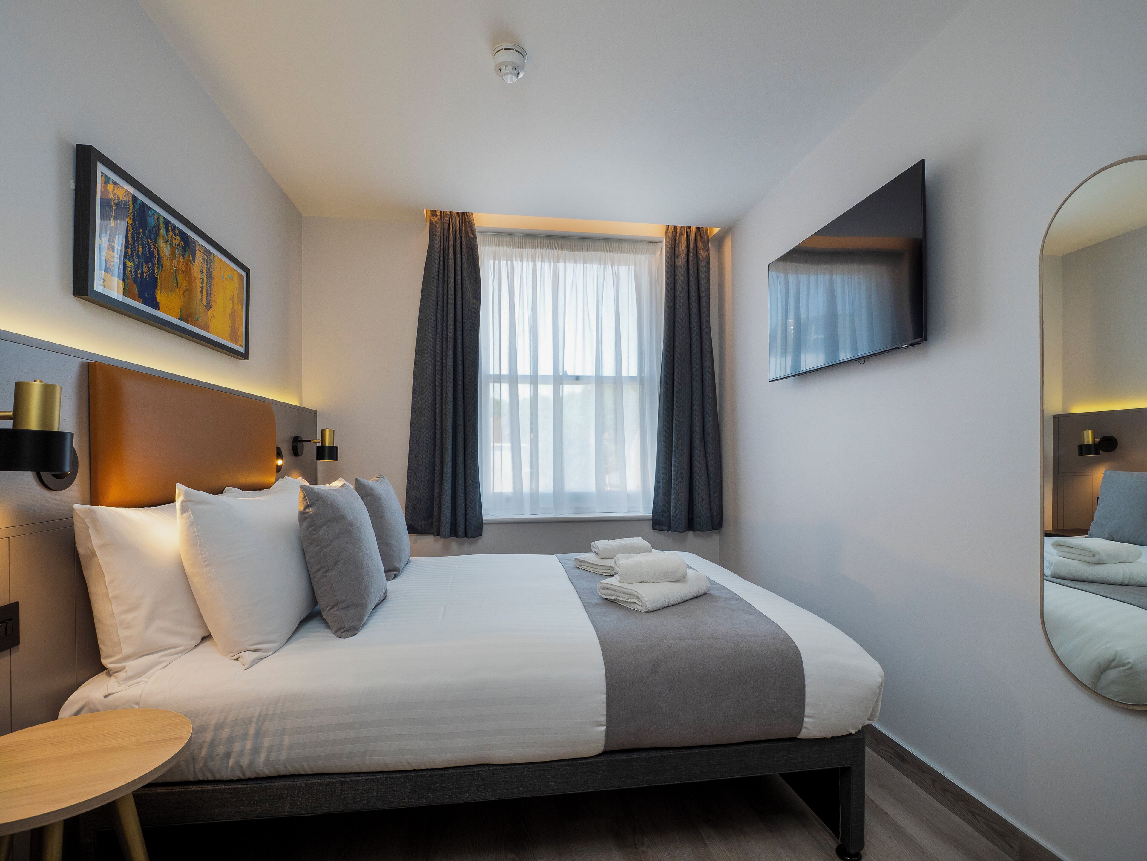 A modern hotel room with a bed, bedside tables, a flat-screen TV, a large window with curtains, and a painting on the wall.