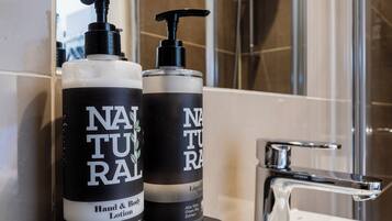 A bathroom with a white sink, two bottles of 'NATURAL RAL' hand and body lotion, and a shower area.