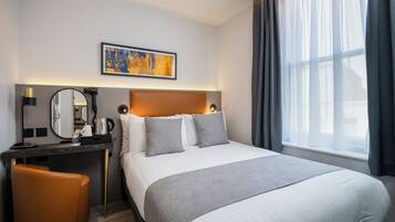 Hotelkamer met een bed, bureau met lamp, stoel, groot raam met gordijnen en een schilderij aan de muur.