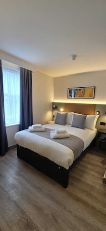 A hotel room with a large bed, a nightstand, a wall-mounted lamp, and a painting above the bed.