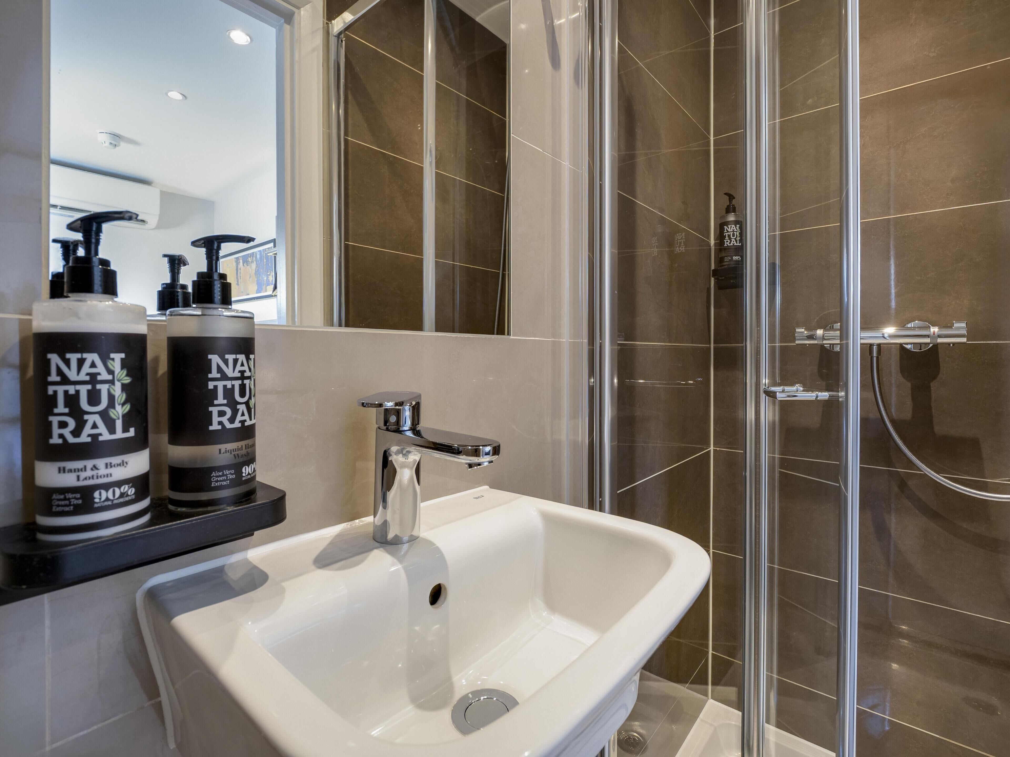 A bathroom with a white sink, a glass shower enclosure, and two bottles of 'NATURAL' hand and body lotion.