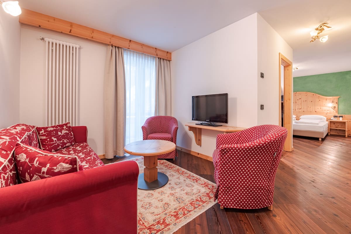 A cozy room with a red sofa, a red armchair, a small round table, a television, and a bed with a wooden headboard.