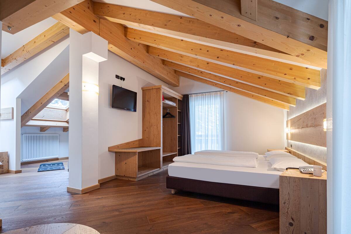 A bedroom with a sloped ceiling, wooden beams, a bed, a bedside table, and a TV mounted on the wall.