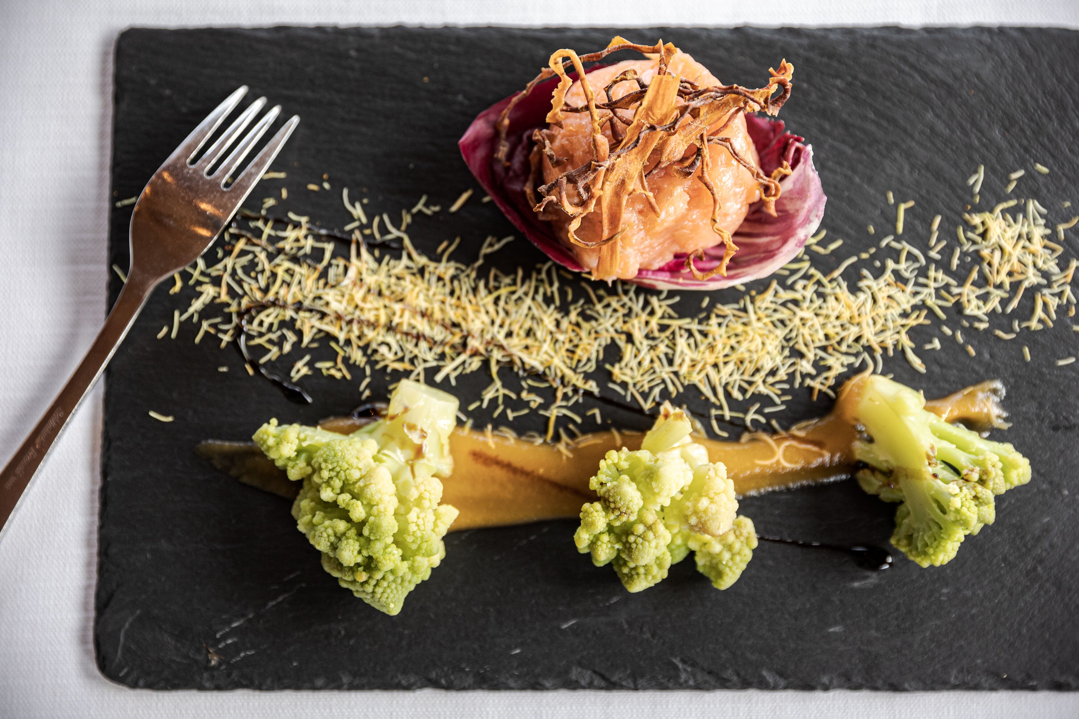 A slate serving board with a dish of food, including broccoli and a red sauce, and a fork on the left.