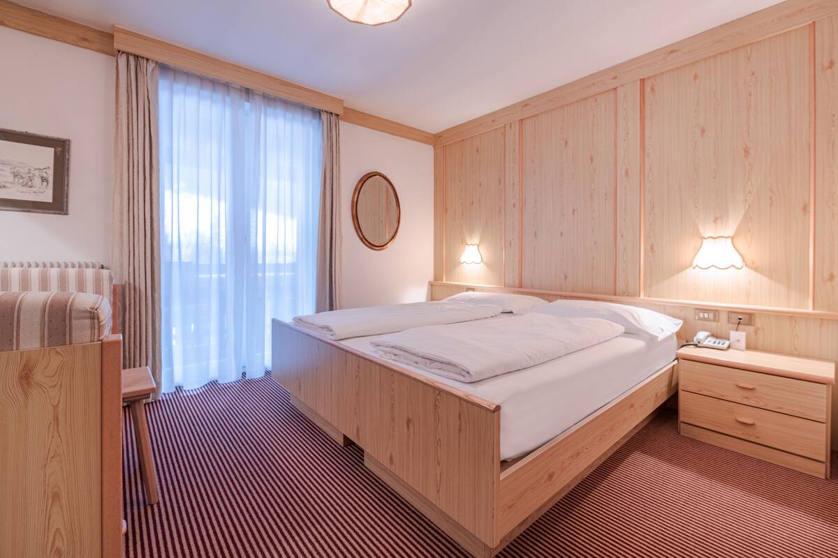 A hotel room with a wooden bed, a bedside table with a lamp, a mirror, and a window with curtains.