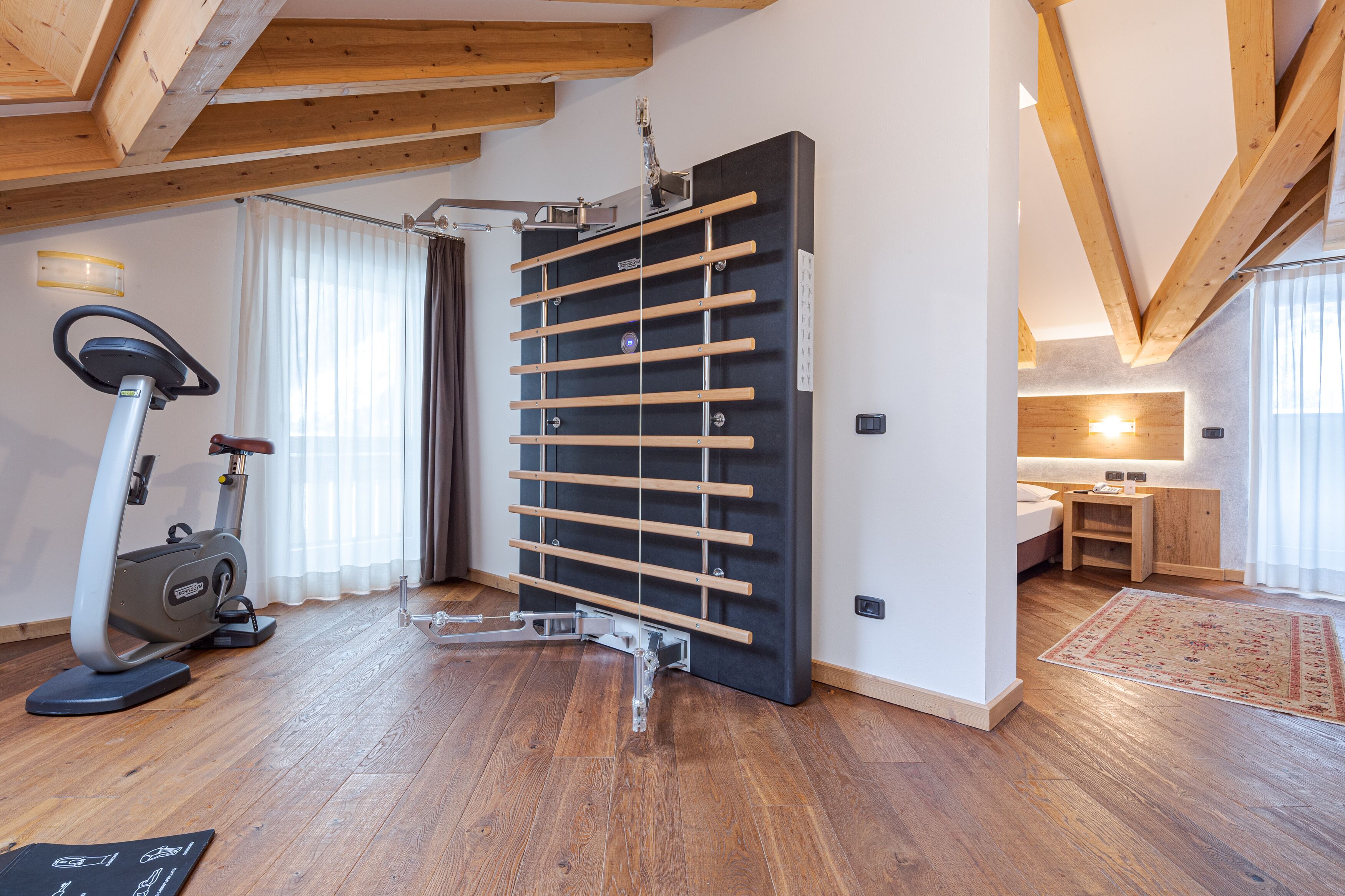 Una habitación con piso de madera, una máquina de ejercicio vertical, una cinta de correr y un amplio ventanal con cortinas.