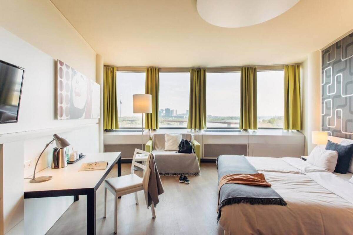 A hotel room with a desk, a bed, a chair, a TV, and a window with curtains.