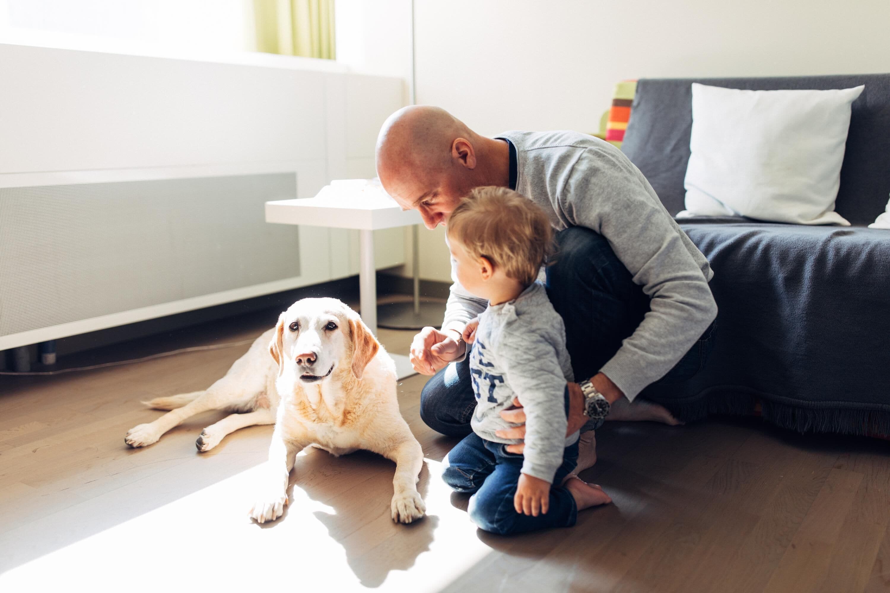 Un hombre y un niño sentados en el suelo con un perro en una habitación con una mesa blanca y un sofá oscuro.