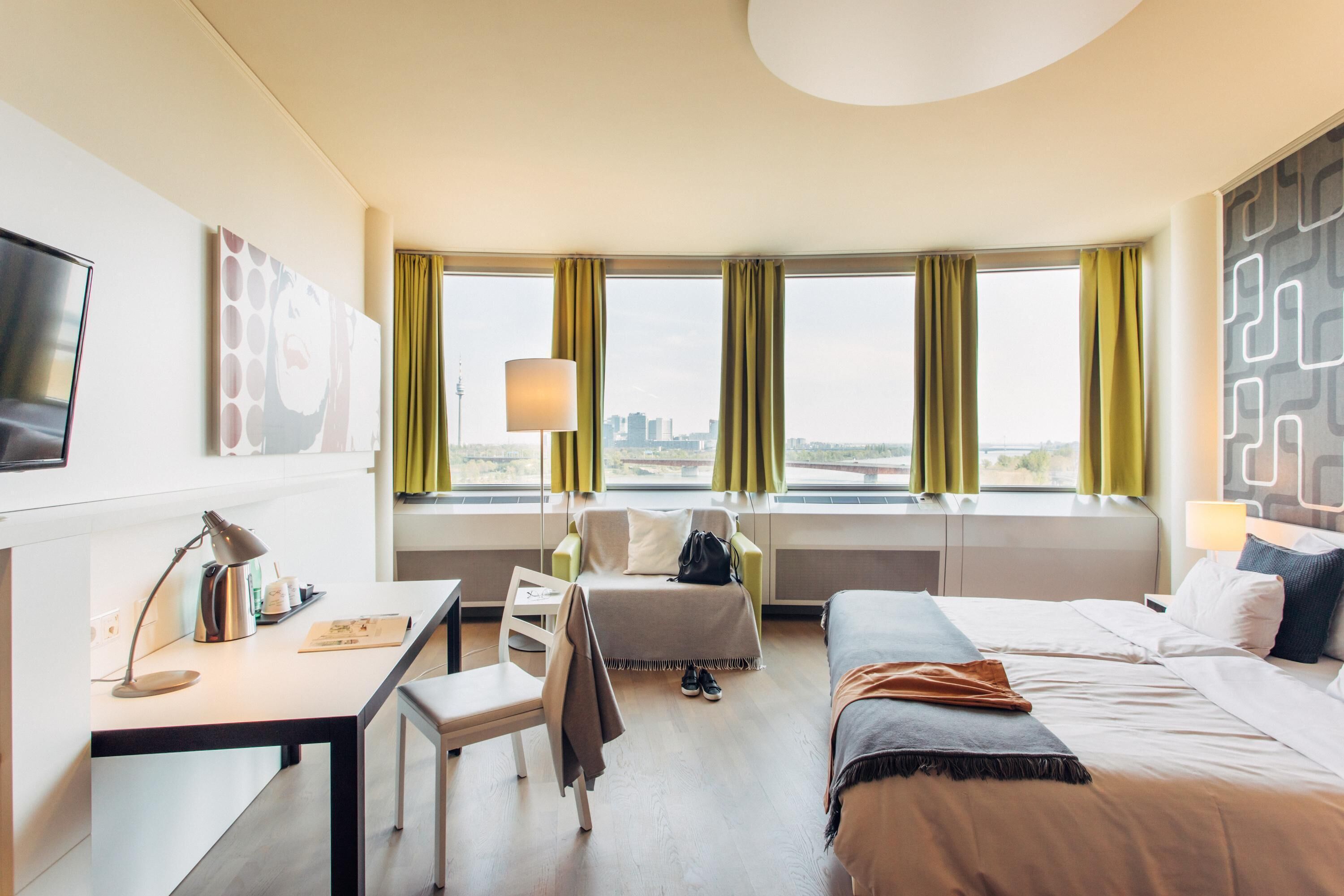 Ein Hotelzimmer mit Schreibtisch, Stuhl, Bett und großen Fenstern mit Vorhängen.
