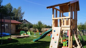 Une structure de jeu en bois, dotée d’un toboggan et d’un mur d’escalade, entourée d’une cour verdoyante et d’arbres.