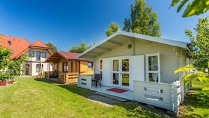 Une maison de style cabane, aux murs blancs et au toit rouge, avec un porche doté d’un paillasson rouge et une petite terrasse.