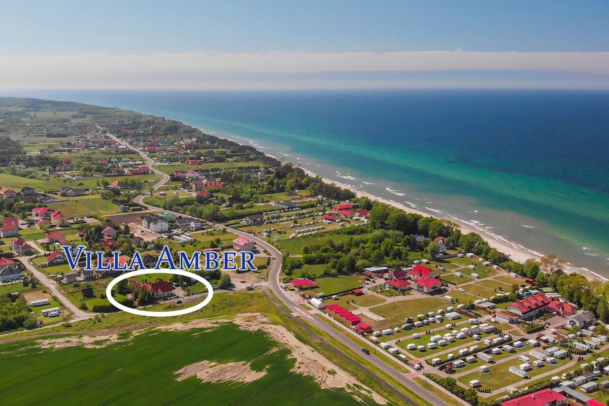 Vista aérea de Villa Amber, con ubicación costera, una carretera que conduce a la playa, campos verdes y varios edificios.