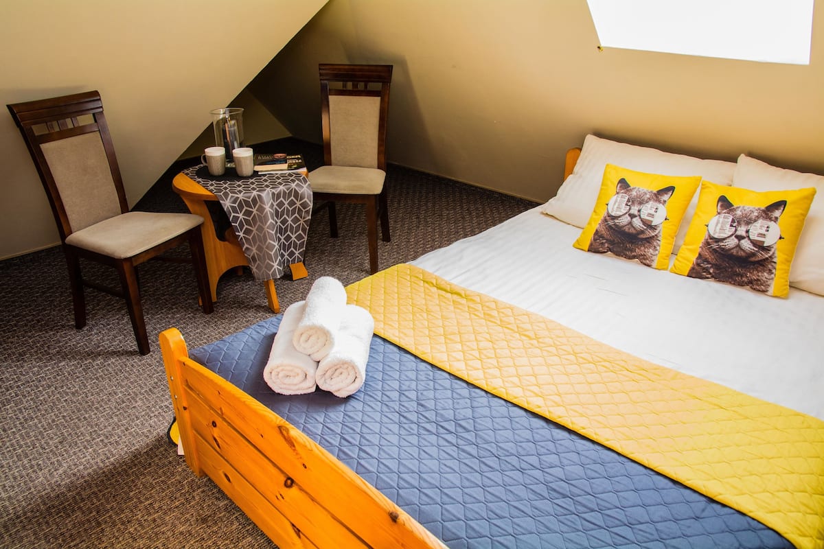 Un dormitorio con una cama, dos sillas, una mesa y una jarra de vidrio, además de un ventanal.