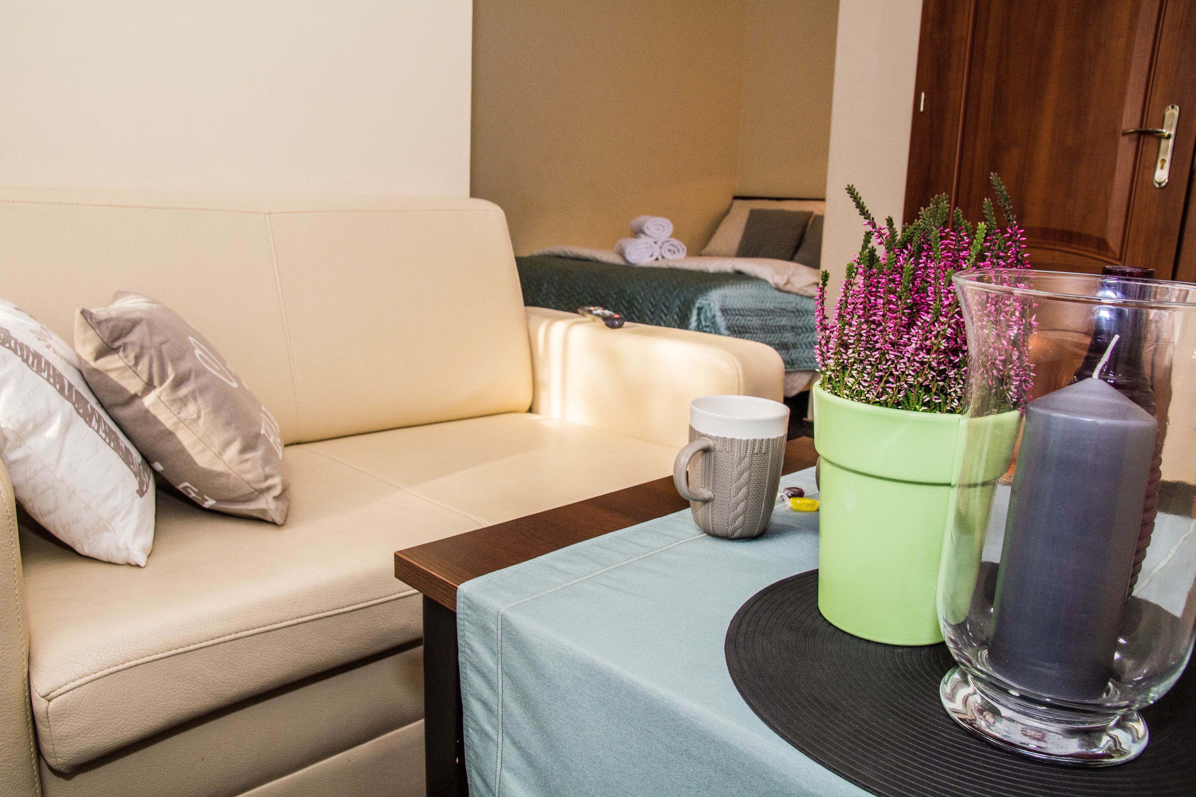 Una habitación con un sofá beige, una planta verde en maceta, un candelabro de cristal transparente y una taza gris sobre una mesa.