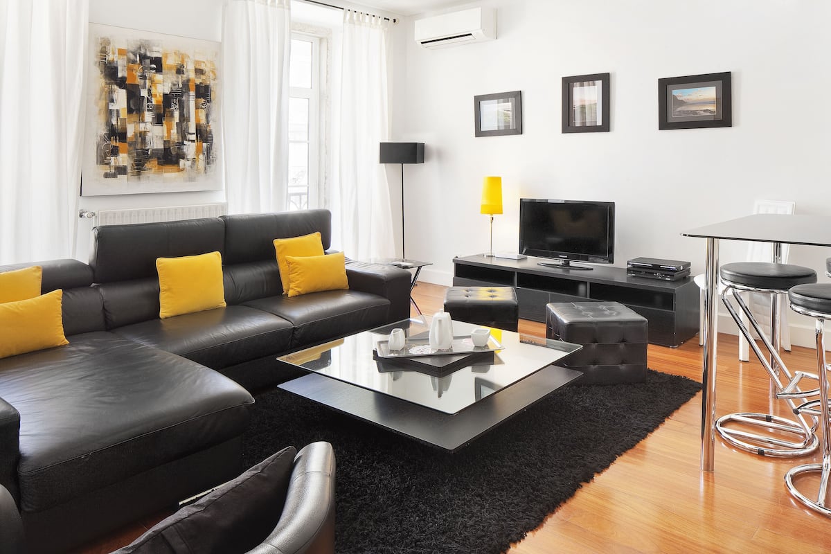 A modern living room with a black leather sofa, a glass coffee table, and a black rug. There is a flat-screen TV, a lamp, and a dining table with bar stools.