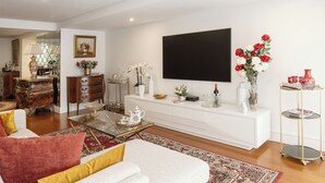 A modern living room with a white sofa, a coffee table, and a TV mounted on the wall. There is a white console table with decorative items and a red flower arrangement. - Lisbon Rentals Chiado (Lisbon)
