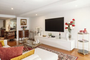 Uma sala de estar moderna com um sofá branco, uma mesa de centro e uma TV fixada na parede. Há um console branco com objetos decorativos e um arranjo de flores vermelhas.