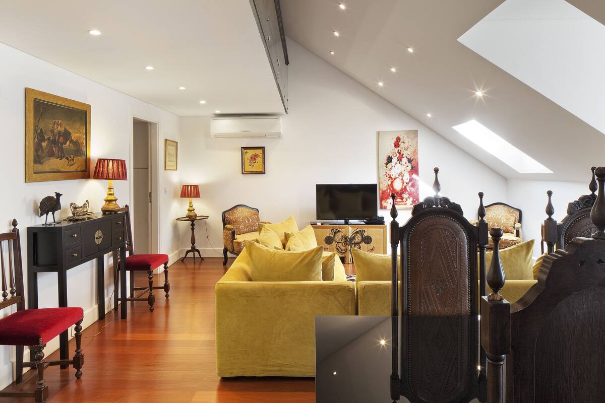 A spacious living area with a high ceiling, a yellow sofa, a television, and a dining area with a table and chairs.