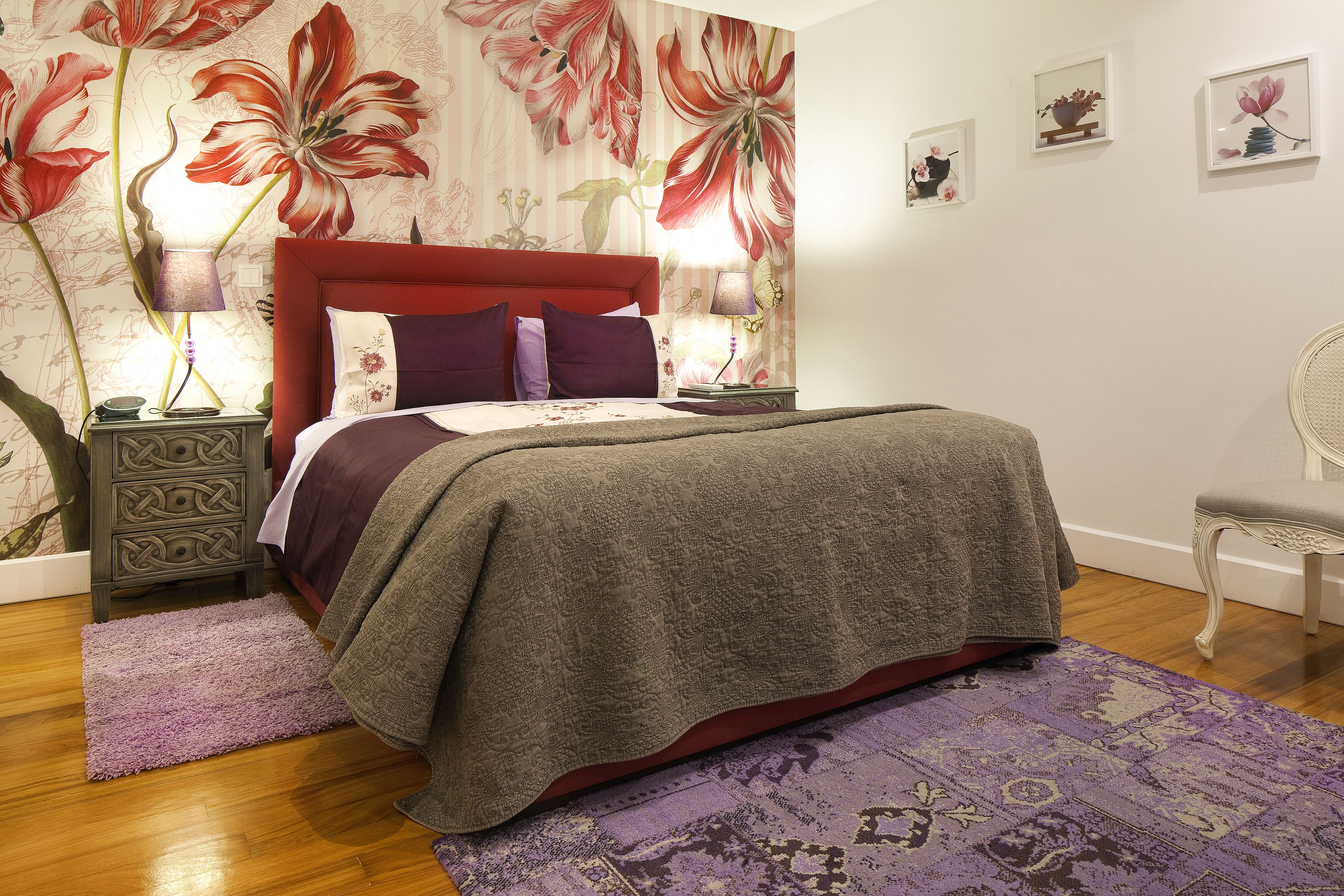 Une chambre à coucher avec une tête de lit rouge, un papier peint à motifs floraux, un tapis violet et une petite table d’appoint avec une lampe.