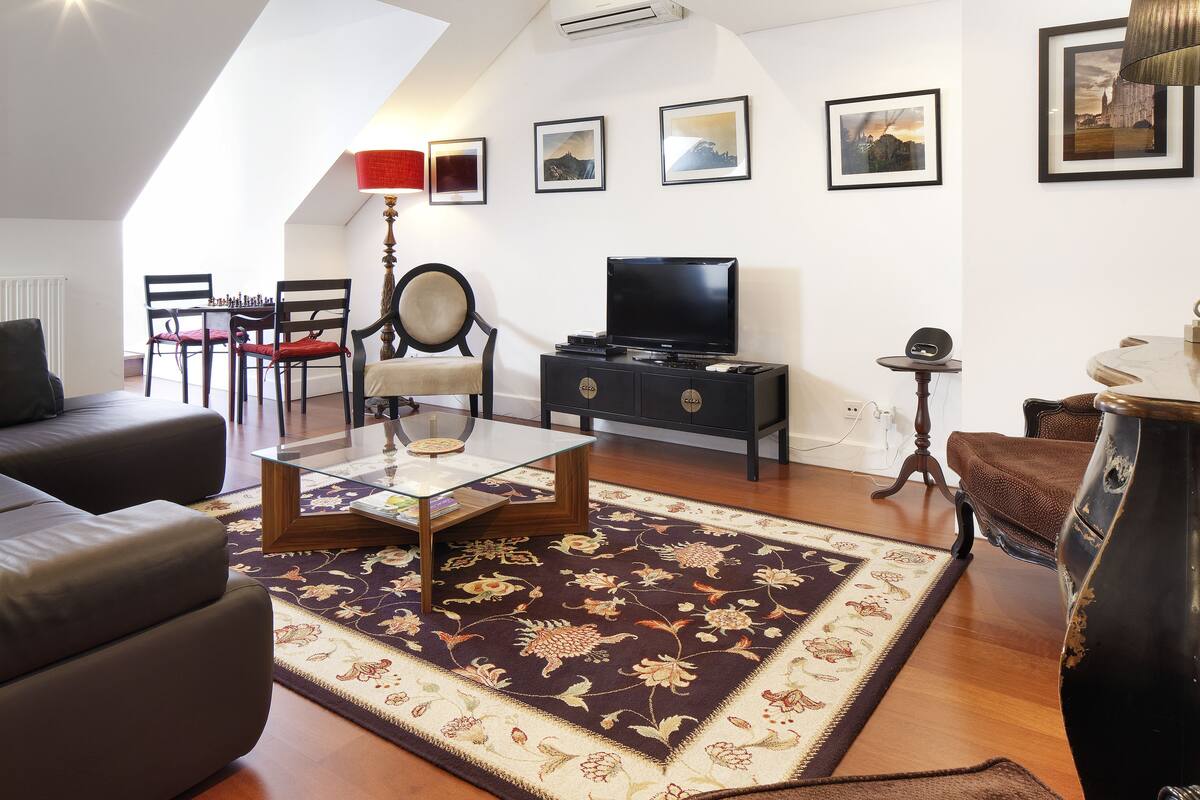A living room with a sofa, a coffee table, a TV, and a red lamp. There are framed pictures on the wall and a carpet with a floral pattern.