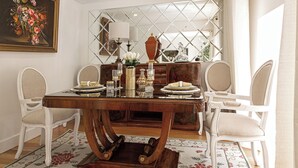 A dining area with a wooden table, white upholstered chairs, a floral rug, and a mirror with a diamond pattern. - Lisbon Rentals Chiado (Lisbon)