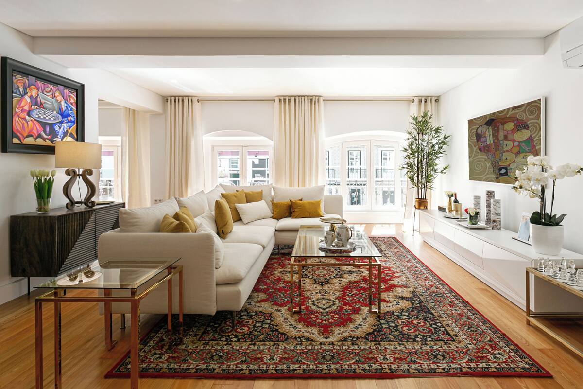 A modern living room with a white sofa, a glass coffee table, and a red patterned rug. There are two windows with curtains, a large painting on the wall, and a potted plant.