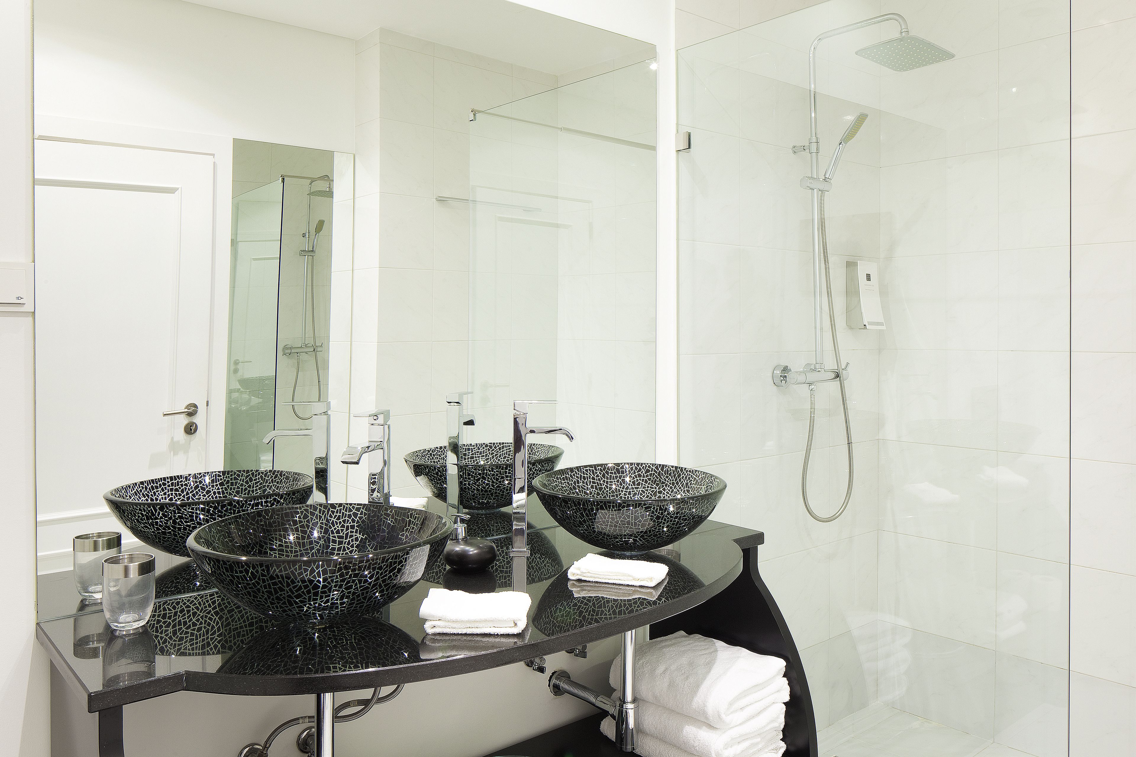 Un baño moderno con ducha de vidrio, lavabo de mármol negro con tres cubetas y tocador con superficie de vidrio.