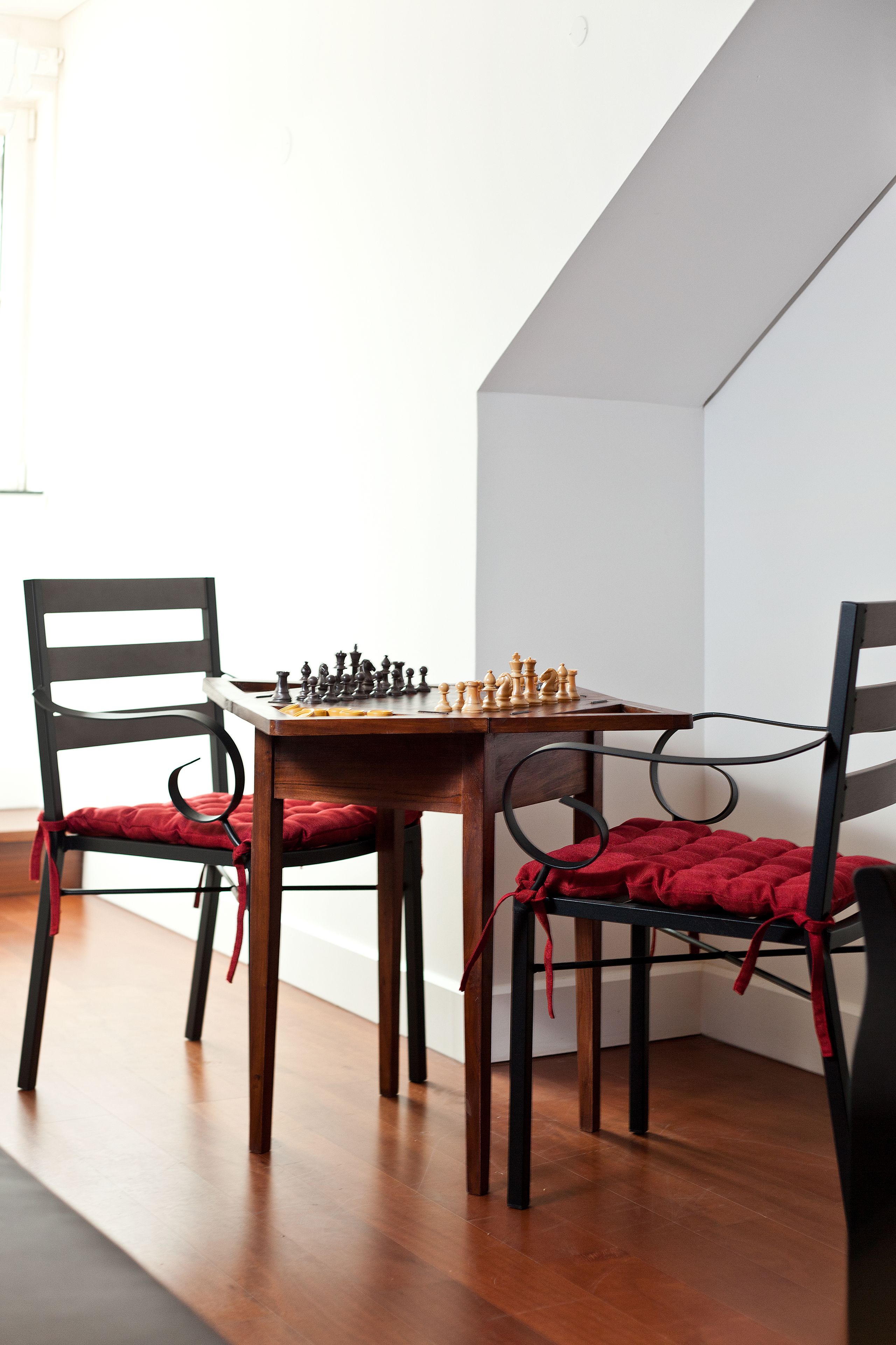 A chessboard set on a wooden table with two chairs, each with a red cushion, in a corner of a room with wooden flooring and white walls.